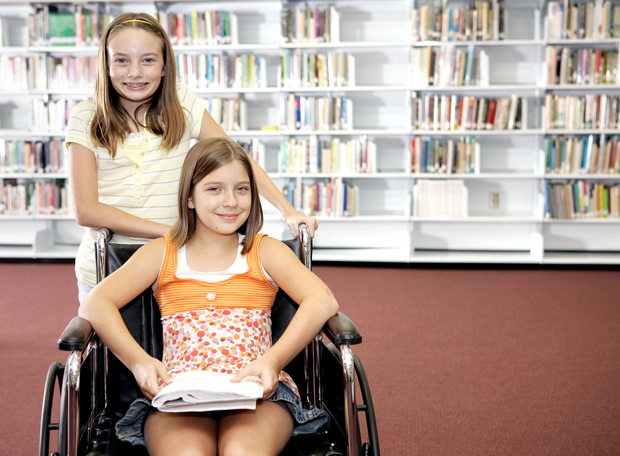 One girl standing behind a girl in a wheelchair, at the library