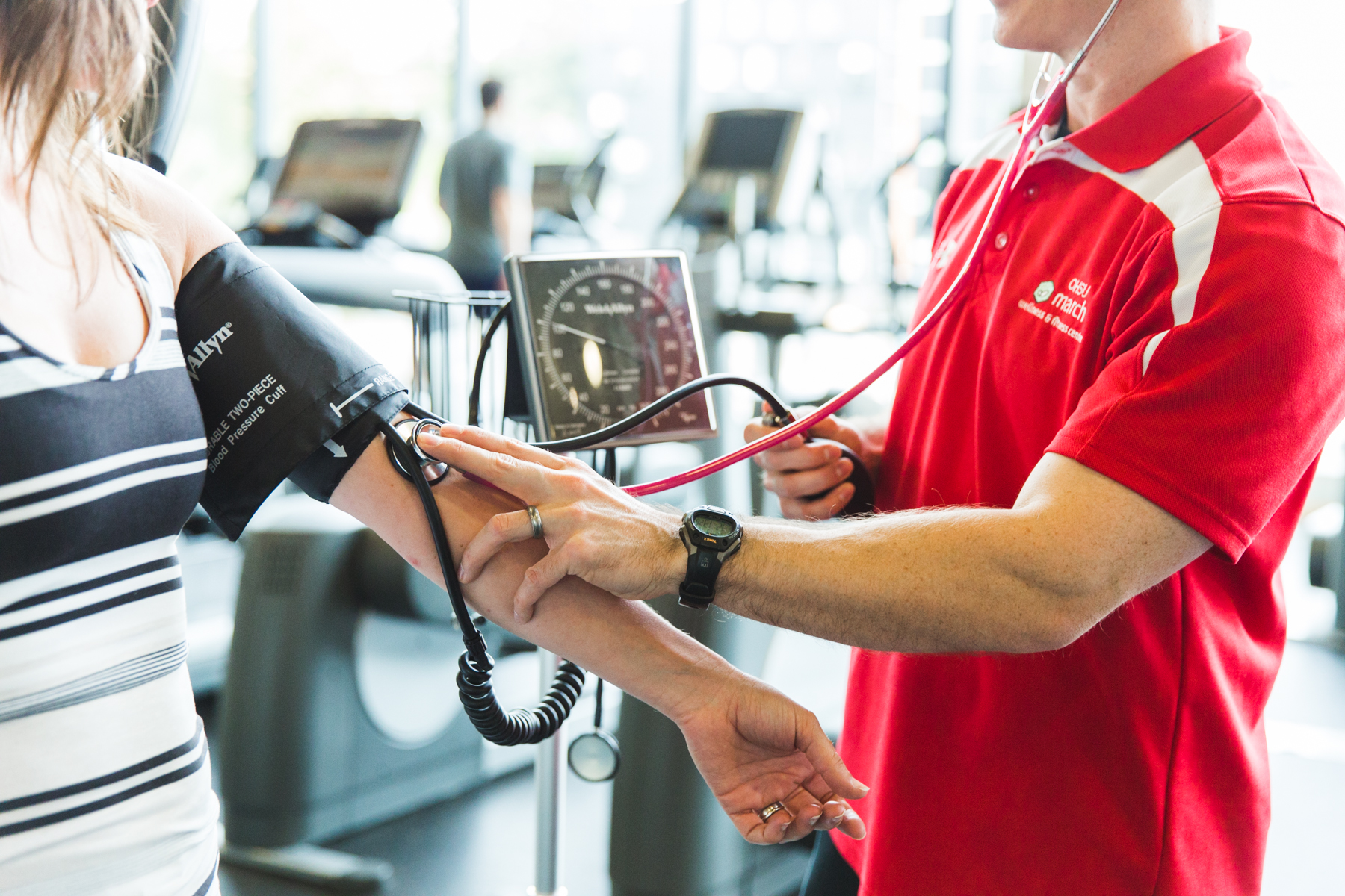 march wellness & fitness center staff member in red polo shirt takes blood pressure of member in black and white striped shirt.