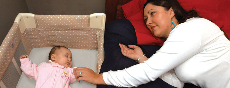 Mother lying down on bed adjacent to baby in crib.