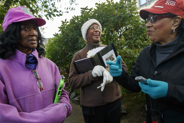 Three women in hats and gloves outdoors, holding a notebook and smart device
