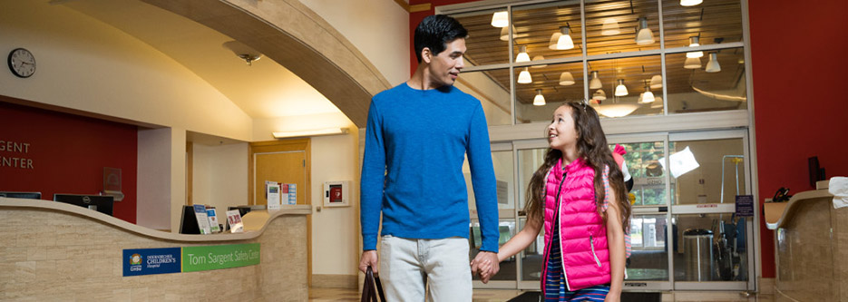 A father and daughter hold hands and smile as they arrive with overnight bags at Doernbecher Children's Hospital.