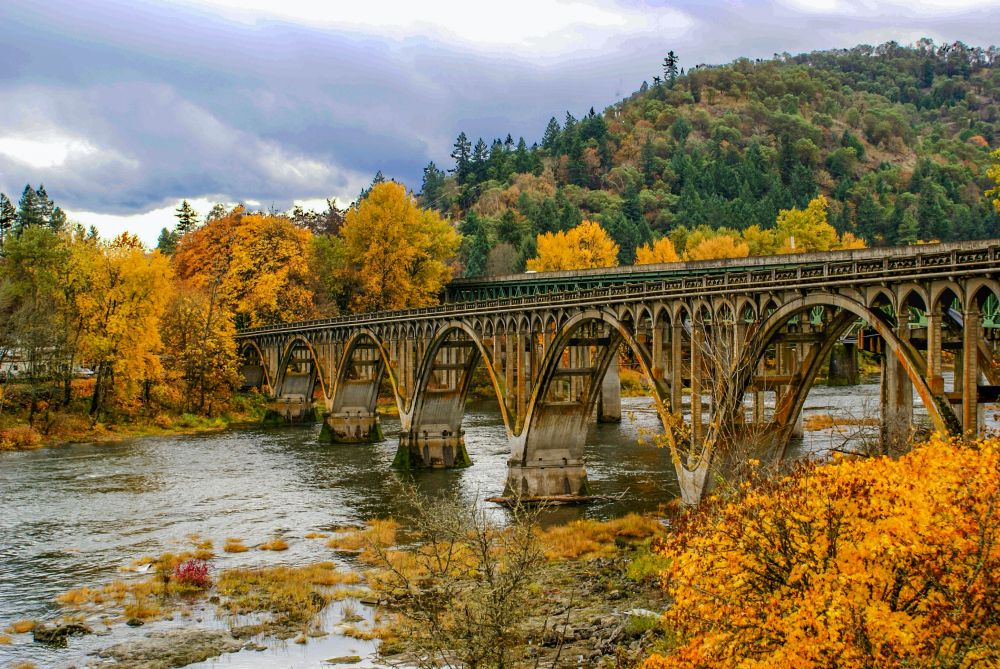 Arch bridge across a river in Oregon in the fall. Foliage on surrounding trees is yellow and orange.