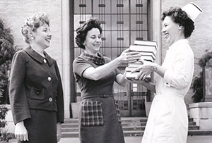 three women stand in front of the 博彩网站 Auditorium, one hands another dressed as nurse a stack of books
