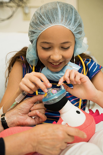 A little girl in a hospital bed pretends to perform a surgical procedure on a stuffed animal.