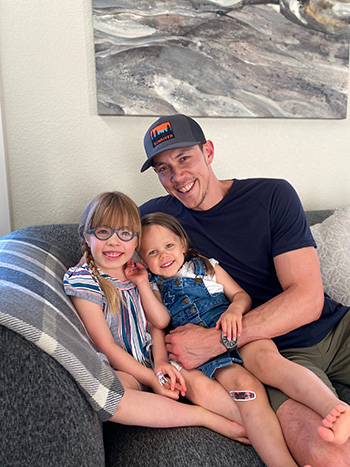 Sienna Balumas with her father and sister at home smiling while sitting on the couch.