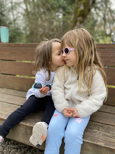 A little girl wearing sunglasses sits on a bench with her sister in a park.