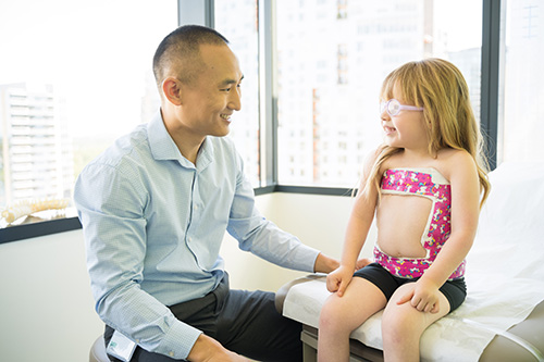 Dr. Scott Yang sitting beside a little girl sitting on an exam table with the cast that helps correct a spinal curve.