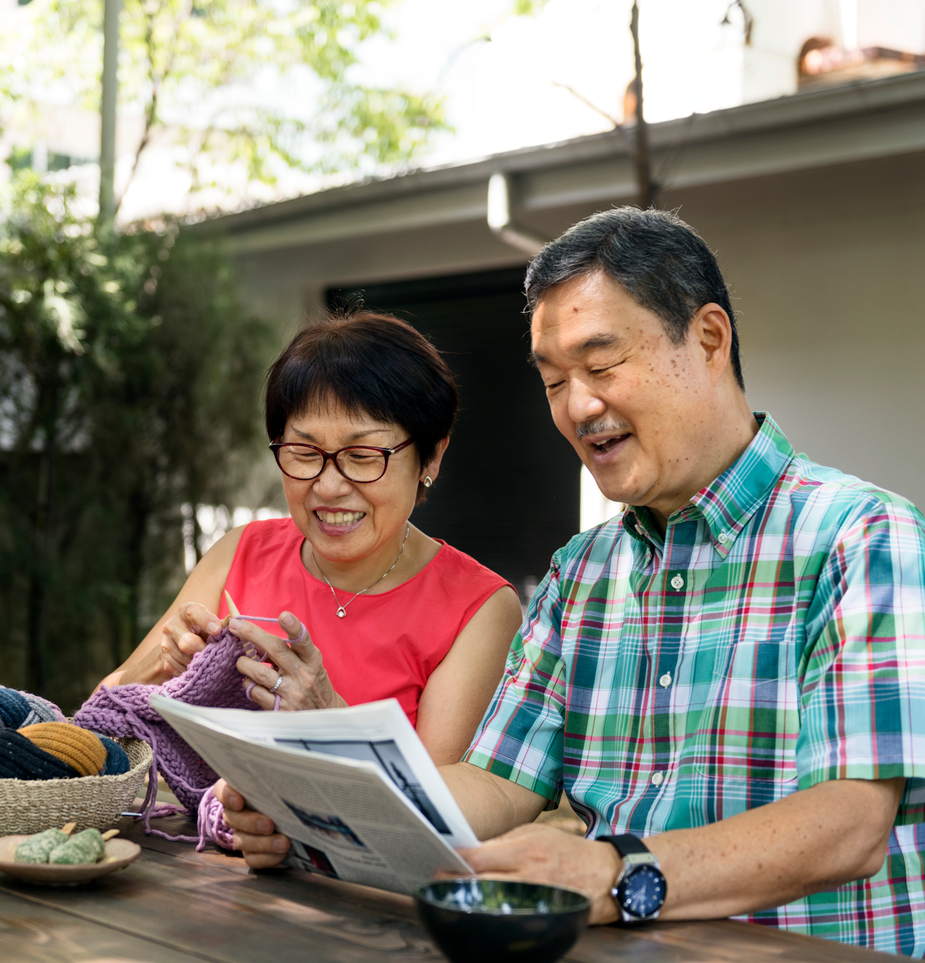 Senior elderly couple sit outside their home knitting and reading newspaper