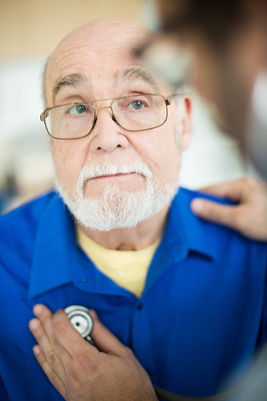 An elderly patient watching as a doctor listens to his heart with a stethoscope.