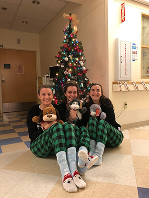 Three female pediatric residents sitting on the ground holding stuffed animals in front of a holiday tree at an 博彩网站 clinic.