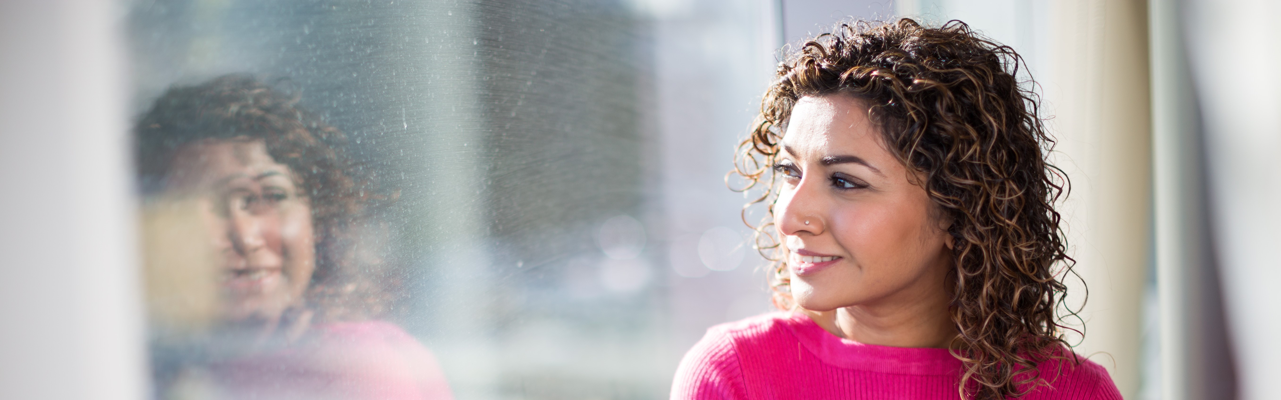 Woman looks out sunny window and her reflection is visible.
