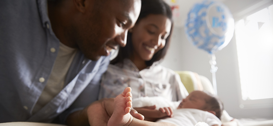 Couple gazes at new baby in hospital with balloon in background. Out of focus except for baby's toes.