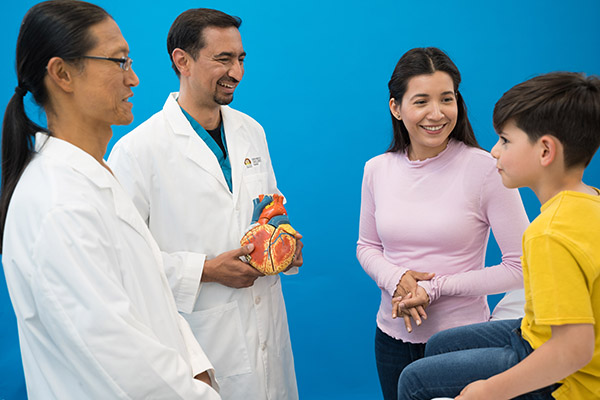 Two male doctors standing and talking to a little boy sitting up on an exam table as his mother stands next to him.