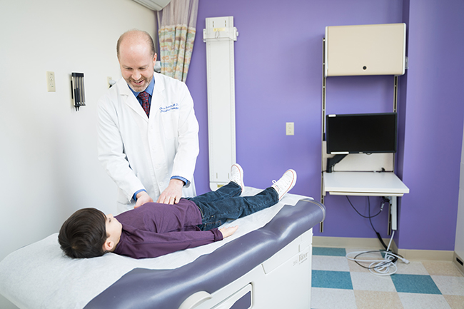 A male doctor feeling the abdomen of a little boy who is on an exam table lying on his back.