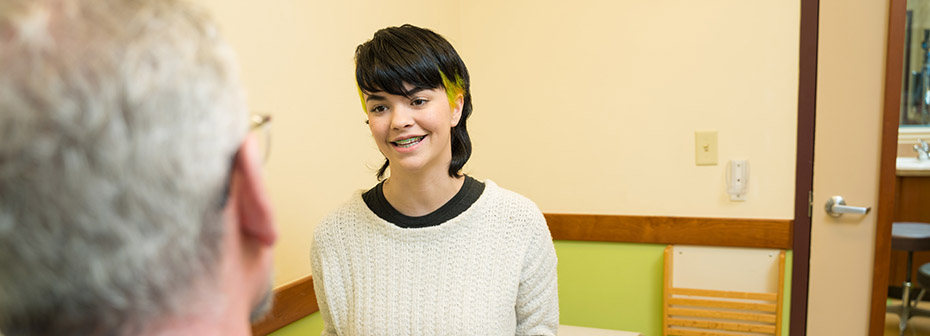 Photo of a young adult patient in an exam room speaking to a healthcare provider.