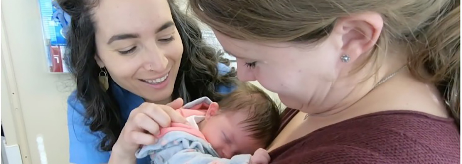 A mother holding her newborn baby, with a female health care provider smiling and admiring the baby.