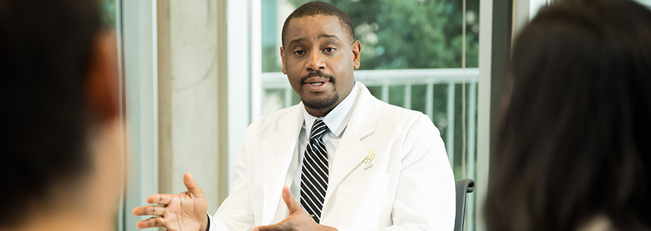 Dr. Luke Masha sitting down at a desk providing an explanation to a patient and his spouse who are sitting across from him.