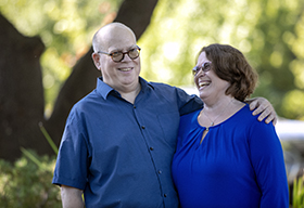 A man with his arm around his wife, both smiling while standing outside.