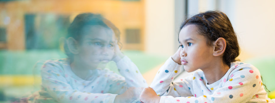 Photo of a girl looking through a window, we see her face and her reflection; she has her hand by her face