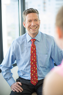 A male doctor sitting down smiling in front of a window while talking to one of his patients, a young girl.