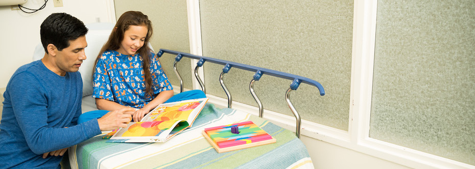 Photo of an adult sitting next to a child who is in a hospital bed, they are looking at books together