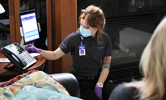 A woman sitting on a couch in her living room watching as a female healthcare provider is showing her how to set up a Hospital at Home visit using a telephone and tablet computer on a side table.