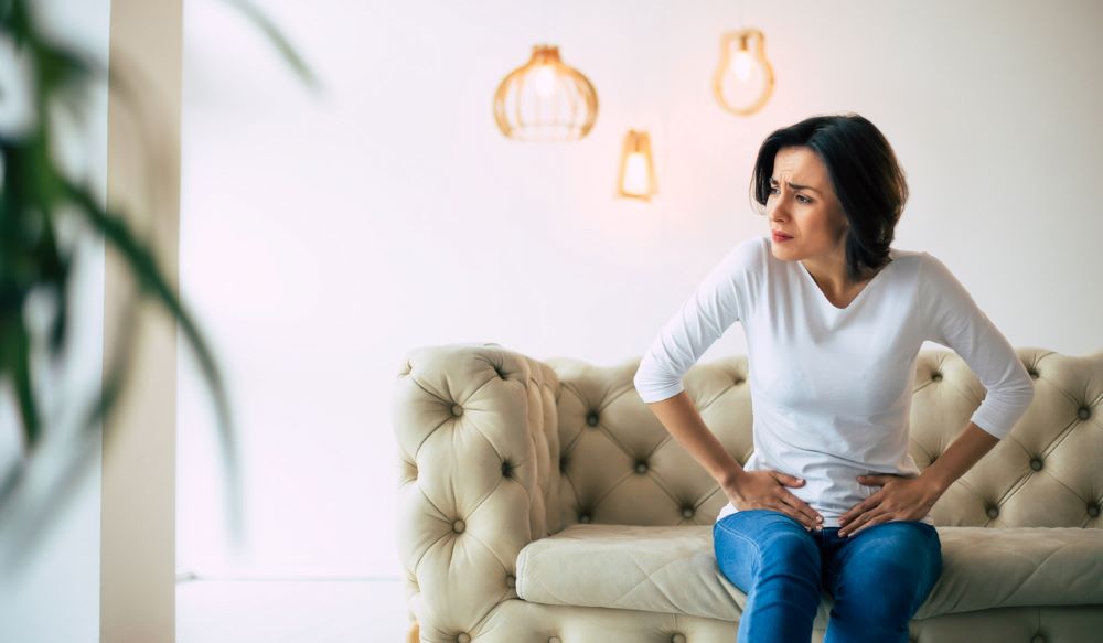 Women crouches over, appearing in pain, on a leather couch