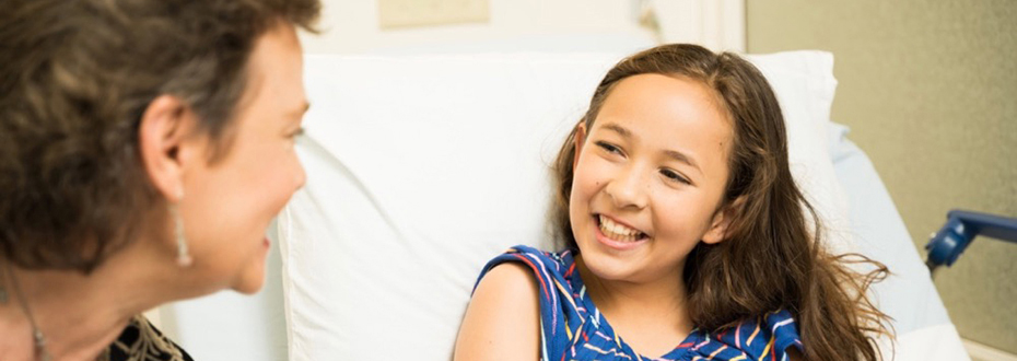 A female care provider talks to a smiling young girl in a hospital bed.