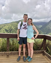 A man and a woman smiling with their arms around each other with green hills in the background.