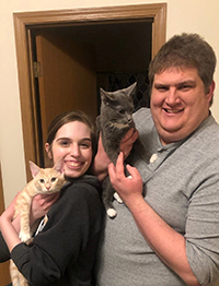 A man and a woman standing in front of a doorway smiling, each holding a cat.
