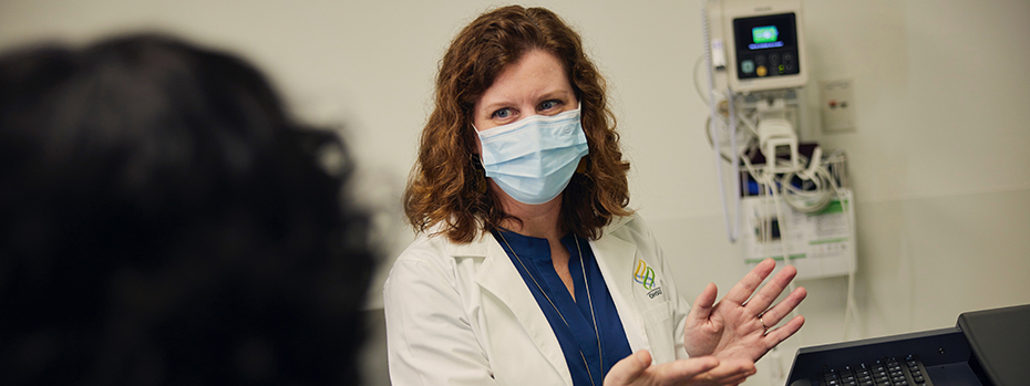 A female doctor wearing a PPE face mask sitting in front of a computer in a hospital exam room explaining something to a female patient.