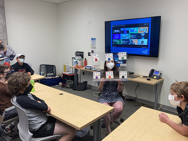 A class of kids at desks wearing PPE masks listening to a woman sitting in the middle of the room giving a presentation and holding a visual aid.