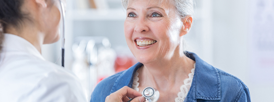 A patient smiles as a care provider examines her with a stethoscope