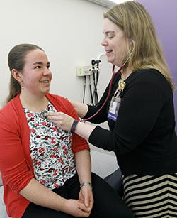 A female doctor stands in front of a woman sitting on a hospital exam table and listens to her heart using a stethoscope.
