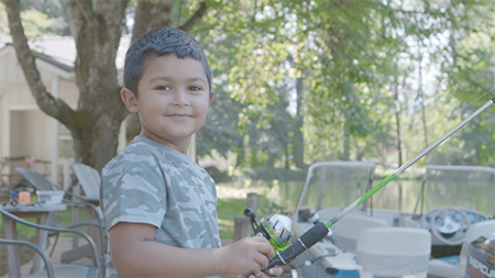 A young boy holding a fishing pole on the shore of a river next to a boat.