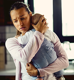A mother with a concerned expression on her face, standing in her kitchen holding her baby.