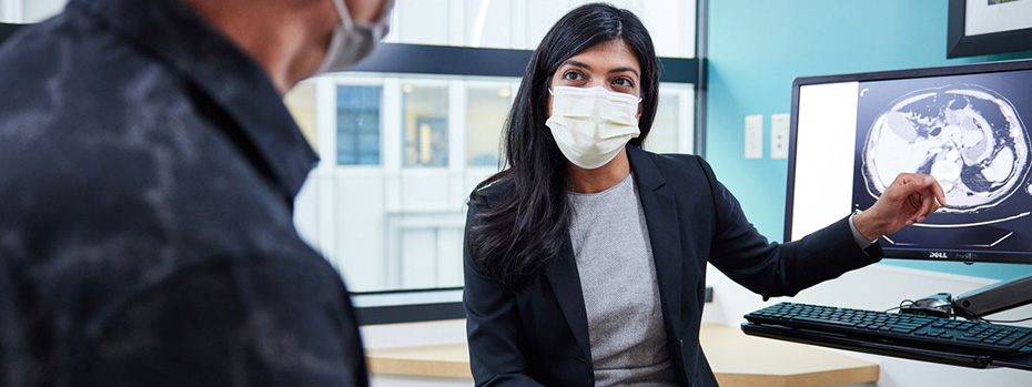 Dr. Divya Sood, reassures a patient in an exam room. She is in the center of the image, pointing to a scan as she explains the HIPEC procedure. The patient is sitting on the left, facing her.