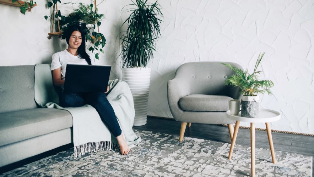Woman does a telehealth visit on laptop from a couch, with plants around her.