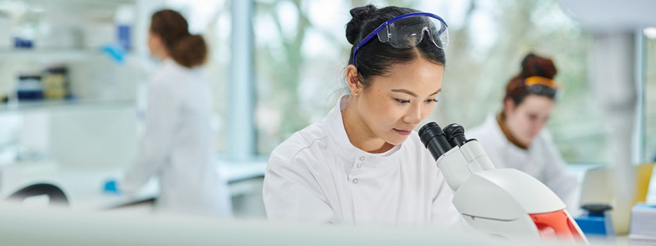 A researcher is peering into a microscope in a busy lab. The mood is light and optimistic.