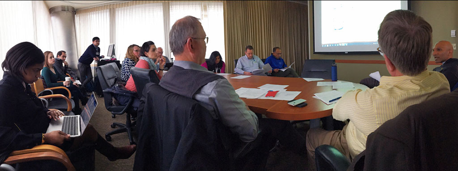 Doctors and researchers crowd around a conference table. They are discussing the best treatment options for a patient. They are consulting notes on laptops and looking at data displayed on an overhead screen.