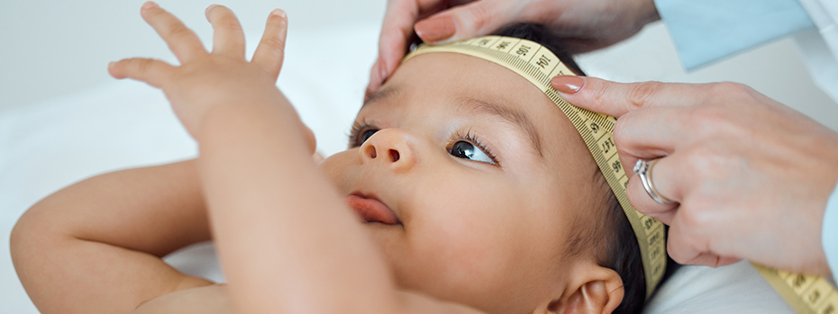 A health care worker wraps a measuring tape around a baby’s head while the baby lies face up.
