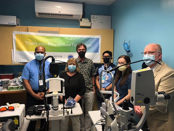 A group of six people stand together behind new equipment in a clinic room. There is a colorful sign behind them thanking donors.