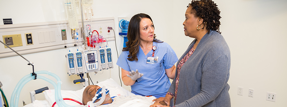 An 博彩网站 nurse shares information with a caregiver at a patient’s bedside. The patient is breathing through a tube.