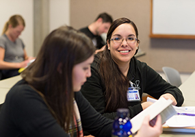 A student with glasses smiles at the camera as she reads from a notebook.
