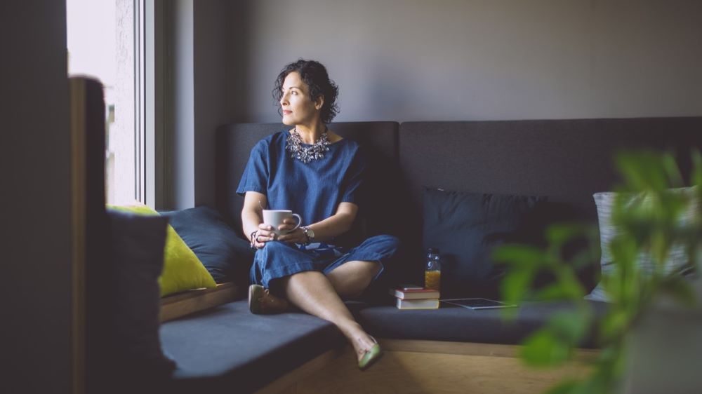 A person in a blue outfit sits sipping a hot beverage while looking out the window.