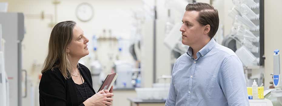 On the left, a woman holds a notebook and speaks to a man on the right. Lab equipment is visible in the background.