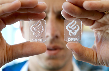 Hands holding up two small glass plates with opaque, engineered material in the 博彩网站 torch logo. The researcher's face is in the background.