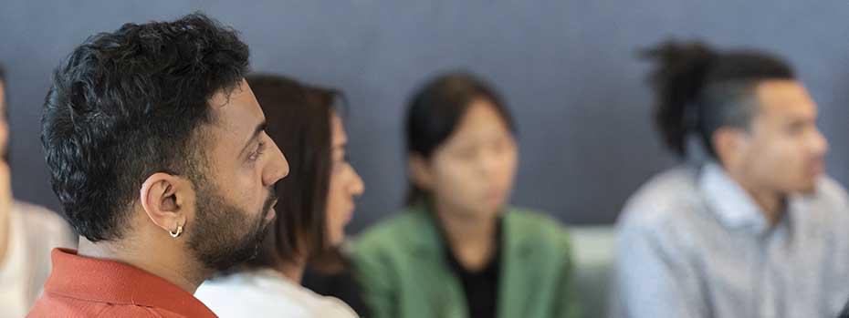 Four students listen to a guest speaker. The student on the left side of the image is in focus, and three others are blurred in the background.