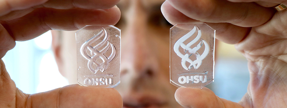 Hands holding up two small glass plates with opaque, engineered material in the 博彩网站 torch logo. The researcher's face is in the background.