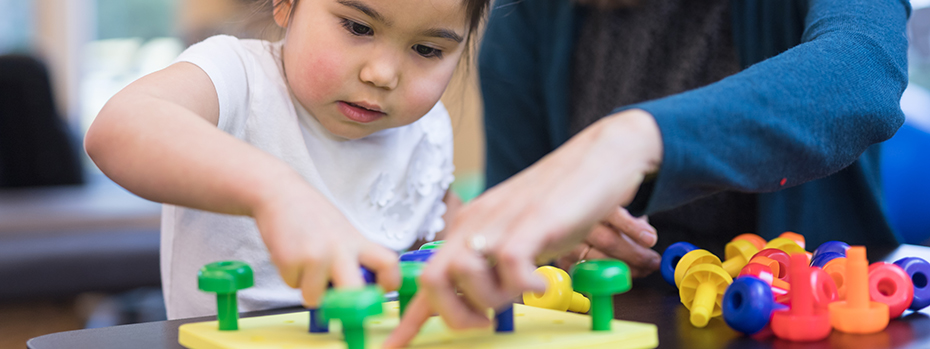 A child places a plastic peg on a board with the help of an occupational therapist pointing out where it goes.